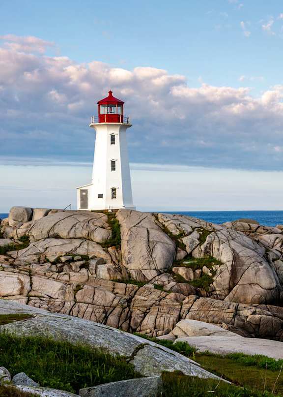 A white lighthouse with a red top stands on a rocky outcrop by the sea.
