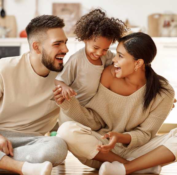 A man and a woman are sitting on the floor with a child, all are smiling and appear to be happy.