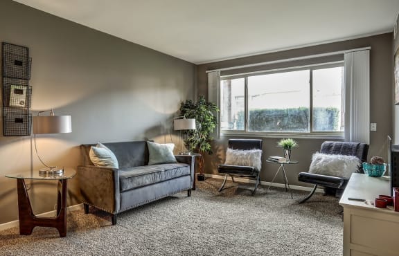 Living room with tan carpet and grey walls. A couch is up against the wall with two chairs under the window. A white dresser is across from the couch.
