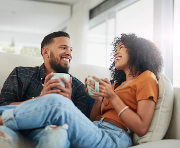 A man and a woman are sitting on a couch, both holding mugs and smiling at each other.