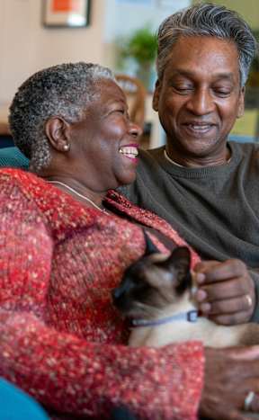 An older couple is sitting on a blue couch with a cat between them.
