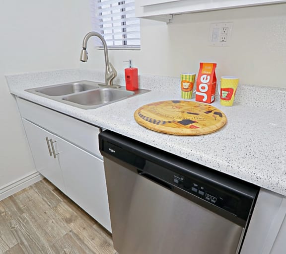 Kitchen countertop area with double sided sink, white cabinetry, and a stainless steel dishwasher at Spring Meadow Apartments in Glendale, Arizona