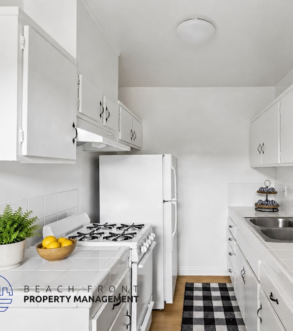 A kitchen with white cabinets and appliances, a black and white checkered mat on the floor, and a logo for Beach Front Property Management in the corner.