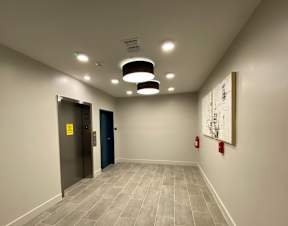 A hallway leading to a waiting area with elevators, wall art, tile-style flooring, and modern circular lights.