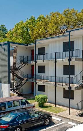 A parking lot with cars, parked outside a three-story building with outdoor staircases.