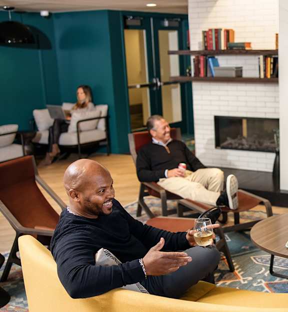 A group of people are sitting around a table in a room with a TV showing a soccer game. at Trio Newton Apartments, Newton, Massachusetts