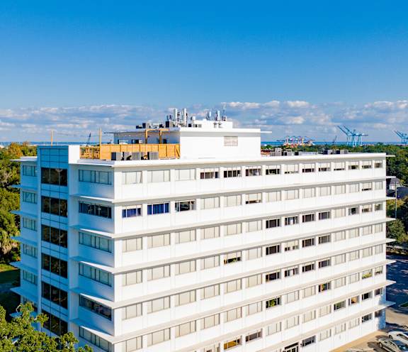A white building with a yellow sign on the top floor is surrounded by trees and other buildings.