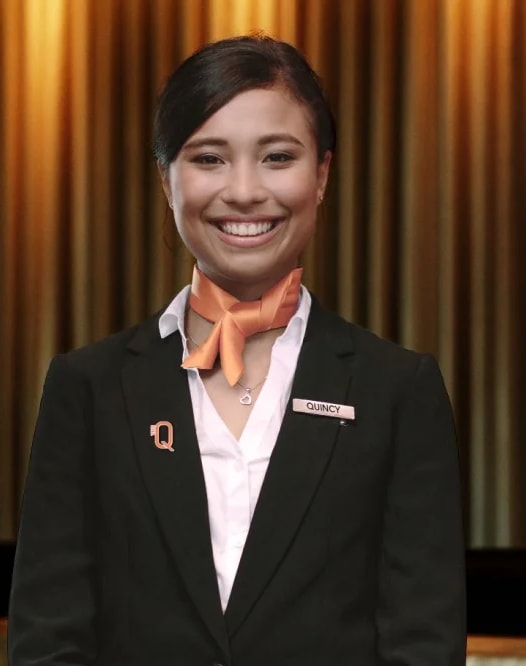A woman in a black suit and orange scarf is standing in front of a red wall.