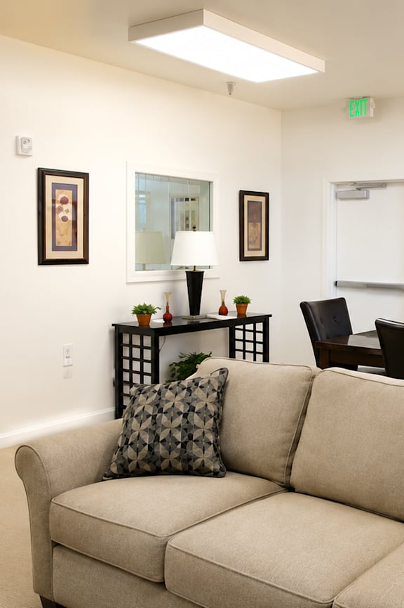 A beige couch with two pillows sits in a room with a white wall and a painting hanging on it.