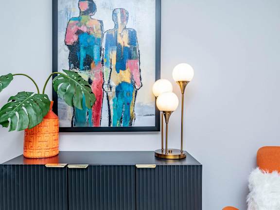 A painting of two people stands behind a black cabinet with a plant, a vase, and a lamp on top.