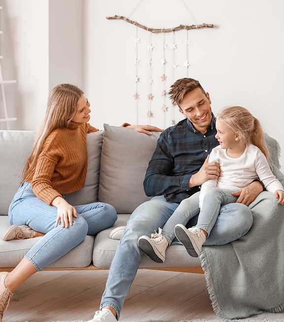 A family of three is sitting on a grey couch in a living room.
