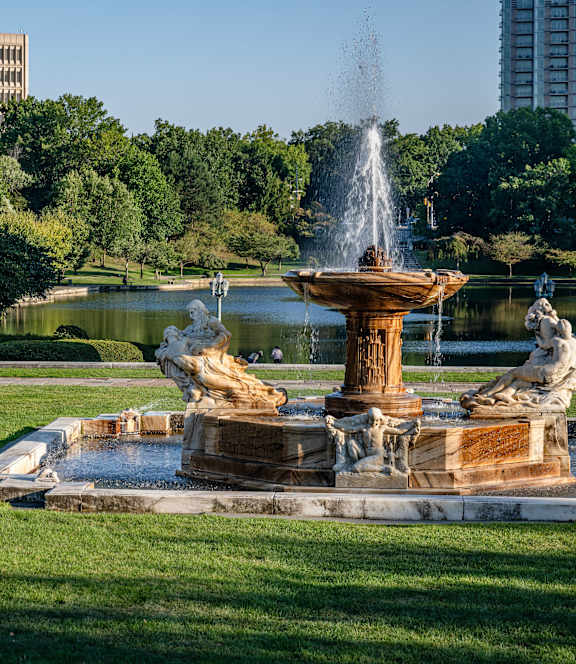 A fountain in the middle of a park with trees and buildings in the background.