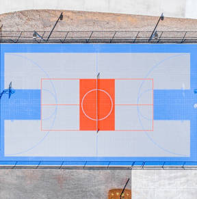 An outdoor basketball court with a blue surface and a red hoop.