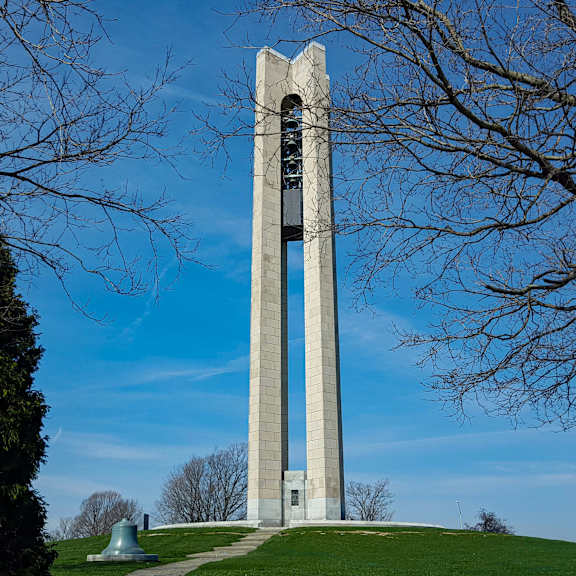 A tall monument stands on a grassy hill with trees in the background.