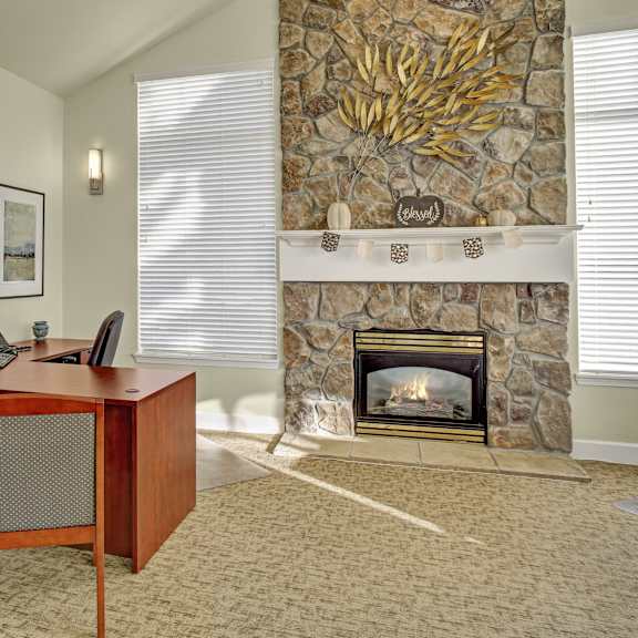 The front office with a wooden desk, chairs, desktop computer, and fireplace surrounded by large windows bringing in natural light at Regency Woods Apartment Homes.