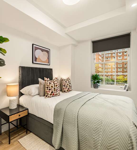 A bedroom with a bed, a nightstand with a lamp, a potted plant, and a window.