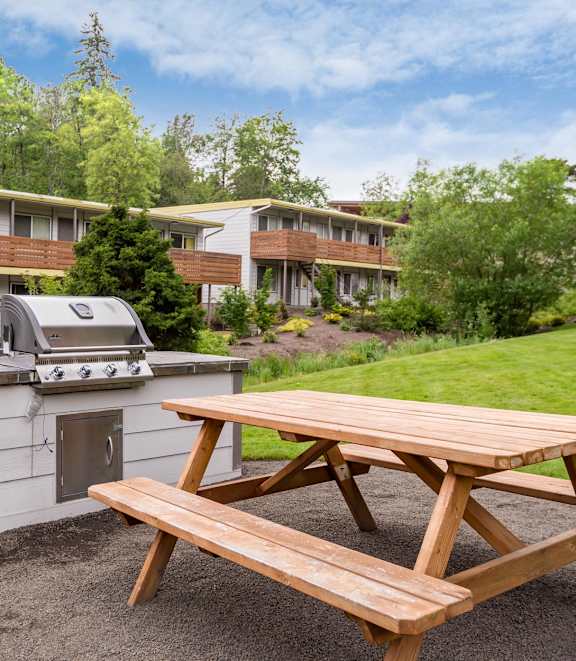 A picnic table is in front of a grill and apartment buildings.