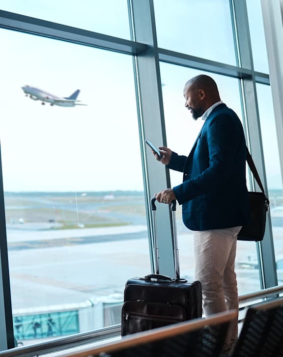 A man in a blue jacket is waiting in an airport terminal with a suitcase, looking out the window at an airplane taking off.