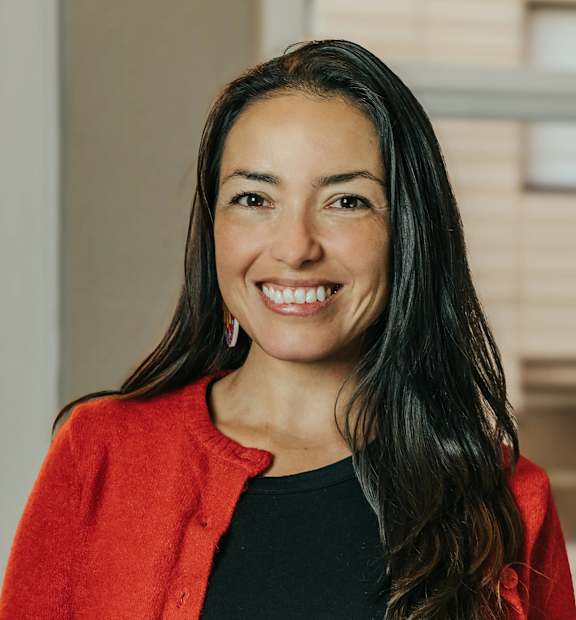 A woman with long dark hair wearing a red cardigan and black top standing in front of a window.