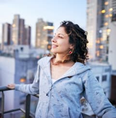 A woman in a blue hoodie stands on a balcony with a cityscape in the background.