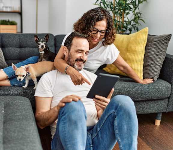 A man and woman are sitting on a couch with a dog on the man's lap, looking at a tablet.