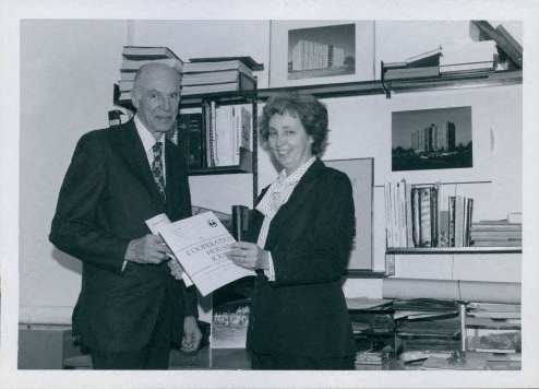 A man and woman in suits stand in an office holding papers.