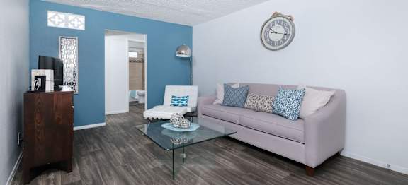 Modern living room in fifteen 50 apartments Las Vegas with grey plank tile floor, grey sofa, glass coffee table, and blue accent wall.