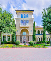A large building with a balcony and arched windows is surrounded by trees and bushes.