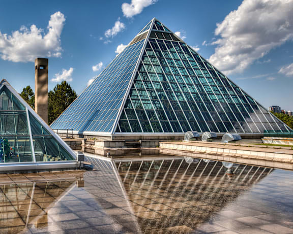 Two pyramid-shaped buildings with glass facades are reflected in a water feature.
