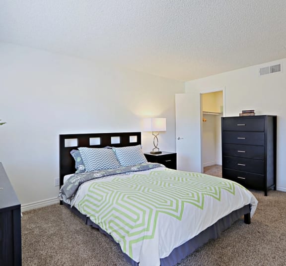 A spacious bedroom with a bed, green patterned bedding, two dressers, and tan carpeting at Spring Meadow Aparmtents in Glendale 85302