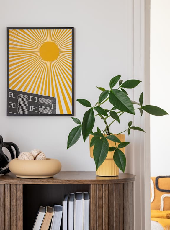 A sunny yellow and black picture hangs on a white wall above a shelf with a plant, books, and sculptures.