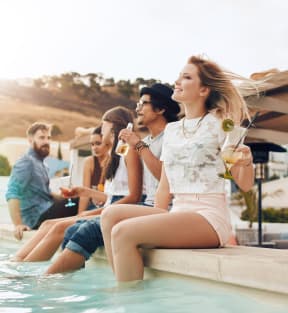 Group of Friends Sitting By Pool Dipping Legs In While Holding Drinks