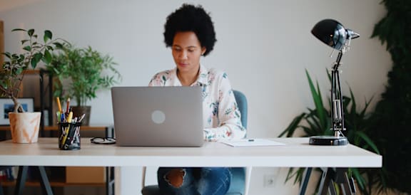 A person is sitting at a desk with a laptop, surrounded by plants and a lamp.