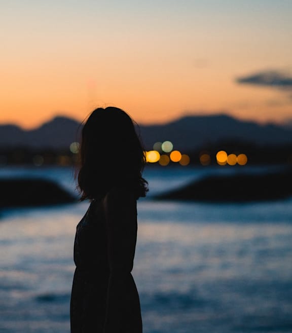 A silhouette of a person stands facing a body of water with a sunset in the background.