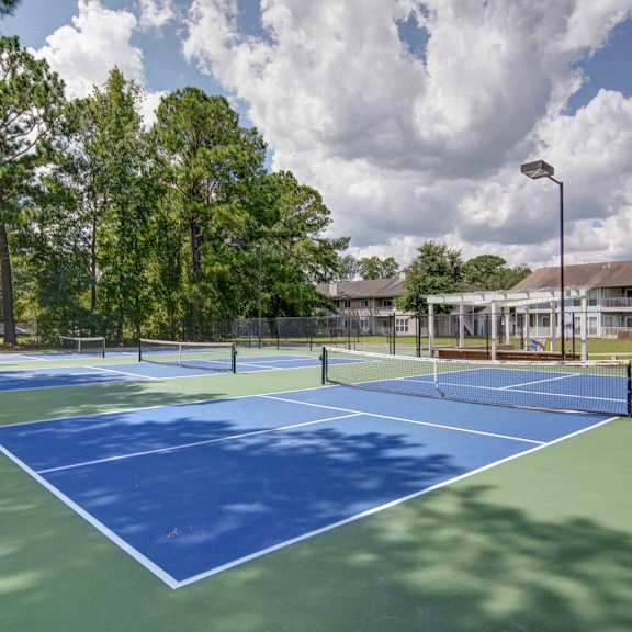 A tennis court with a blue surface and white lines is surrounded by trees and apartment buildings.