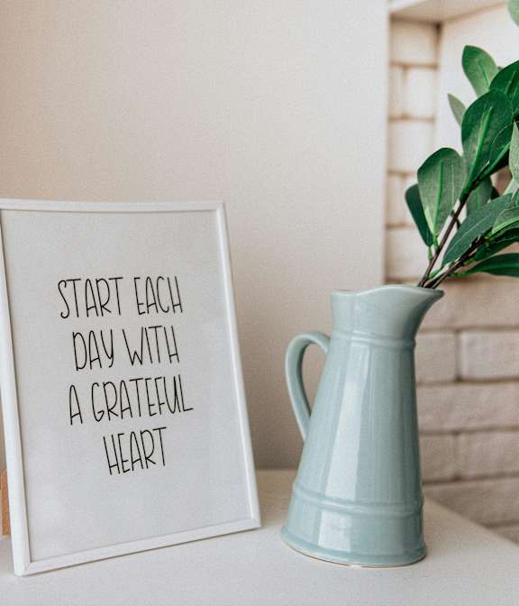 A white framed sign that says "Start each day with a grateful heart" sits on a table next to a blue pitcher.