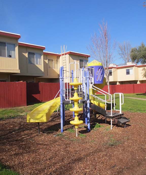 American River Gardens playground on bark with grass & concrete surrounding, in front of apartment buildings with red fencing at American River Gardens, Sacramento, California