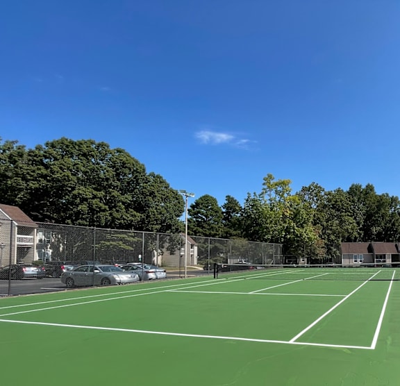 A tennis court with a green surface and white lines, surrounded by a chain link fence. at Lakes Edge Apartments, Apartments, Greensboro, NC