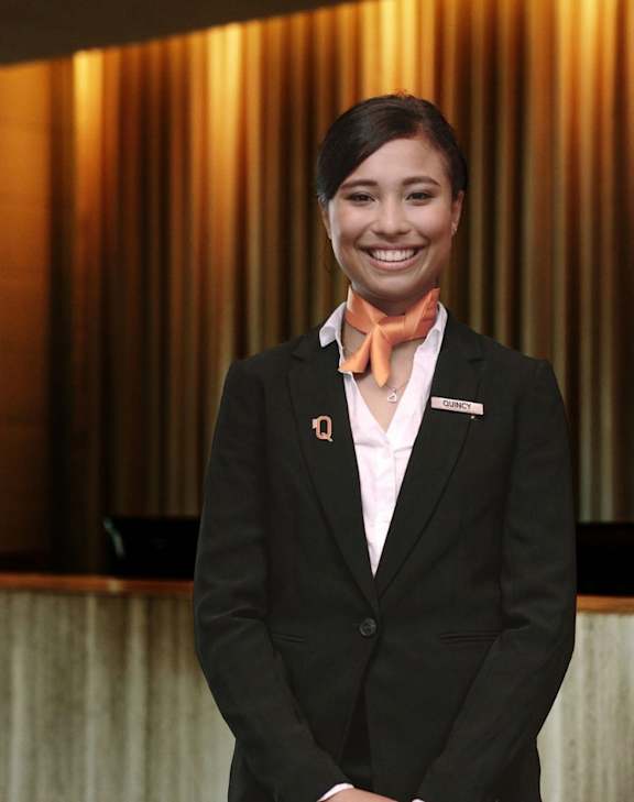 A woman in a black suit and orange scarf is standing in front of a red wall.
