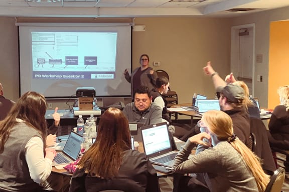 a group of people sitting around a table with laptops in front of a projector screen