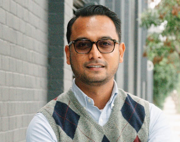 A man wearing glasses and a sweater vest stands with his arms crossed.