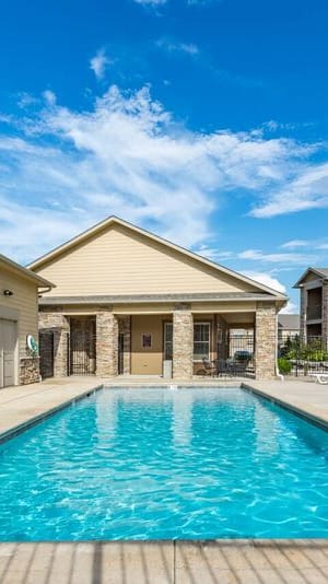 1-Bedroom Apartments In Lawrence, KS - Camson Place - A Large Rectangular Community Swimming Pool Surrounded By Tan Lounge Chairs And Stone-Accented Buildings Under A Blue Sky.