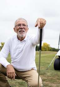 A man is squatting and holding a golf club in front of a golf cart.