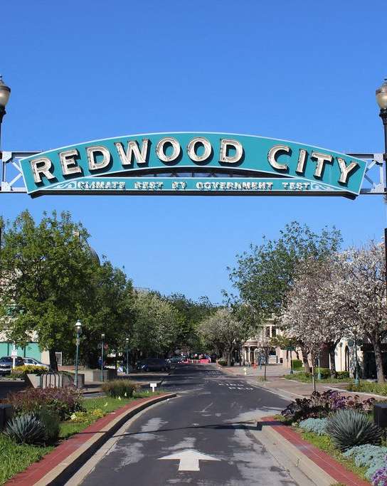 A street sign for Redwood City stands in the middle of a tree-lined street.