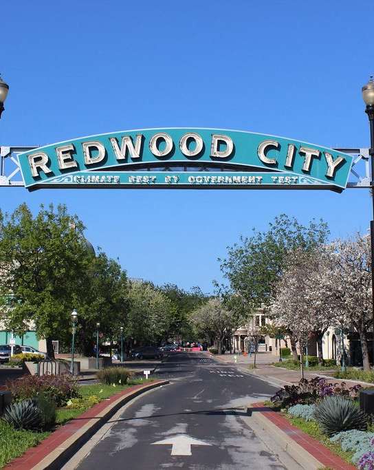 A street sign for Redwood City stands in the middle of a tree-lined street.