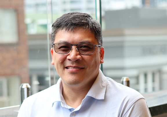 A woman in a grey suit stands in front of a framed picture of a boat.