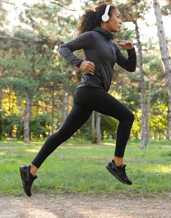 A woman in black running gear is running on a dirt path in a wooded area.