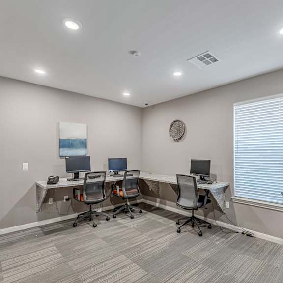 A room with a carpeted floor and a white ceiling with lights, containing a desk with a laptop and a monitor, a chair, and a wall clock.