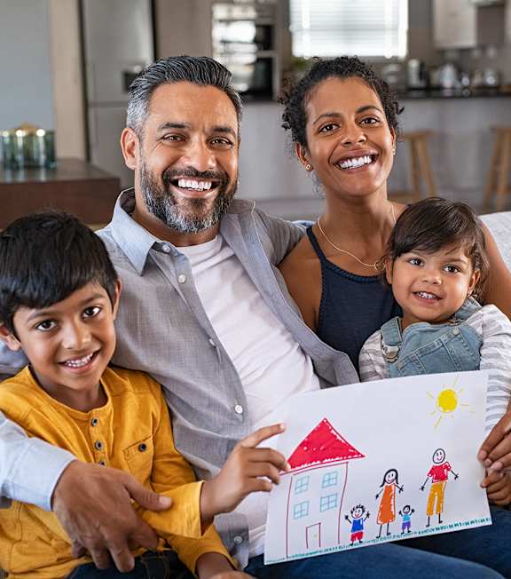 A family of four is sitting on a couch, with a child holding a drawing of a house.