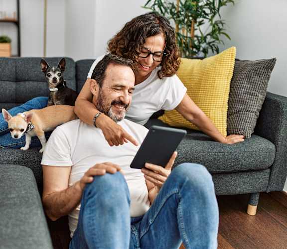A man and woman are sitting on a couch with a dog on the man's lap, looking at a tablet.